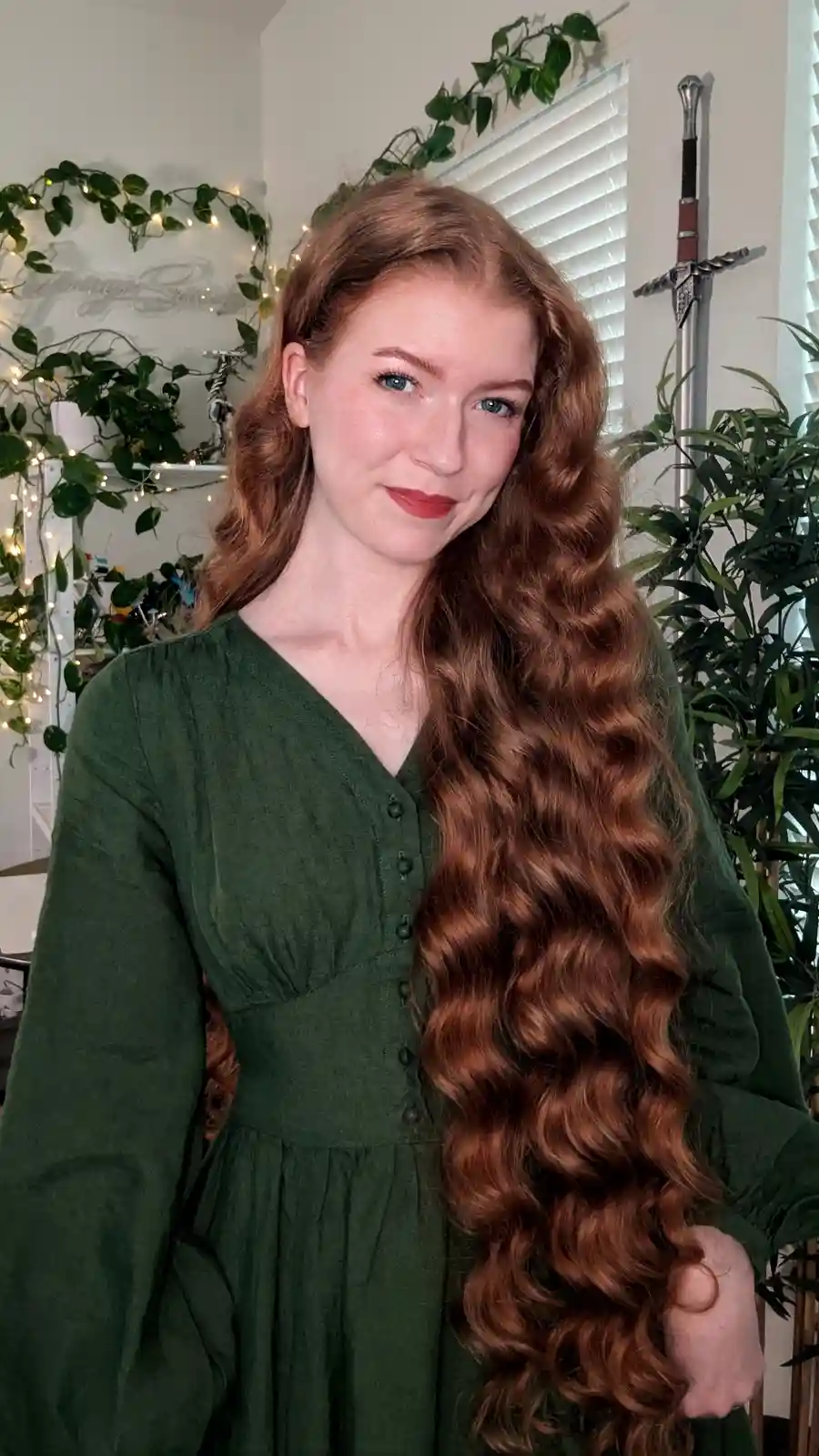A woman with long, wavy auburn hair and blue eyes wears a green dress and smiles at the camera. Behind her, a sword is…