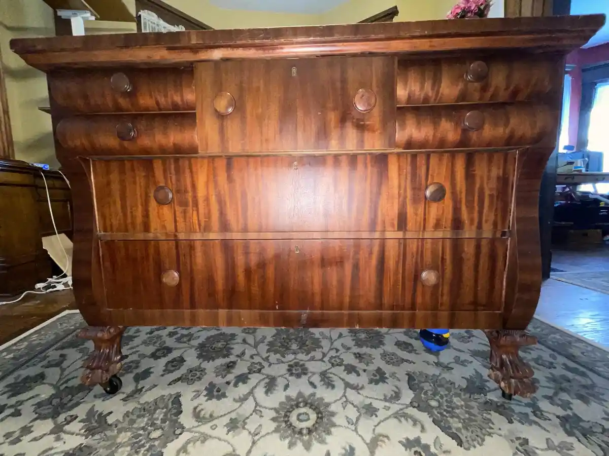 A large, ornate wooden dresser with multiple drawers and carved feet stands on a patterned rug. The dresser has a glossy…