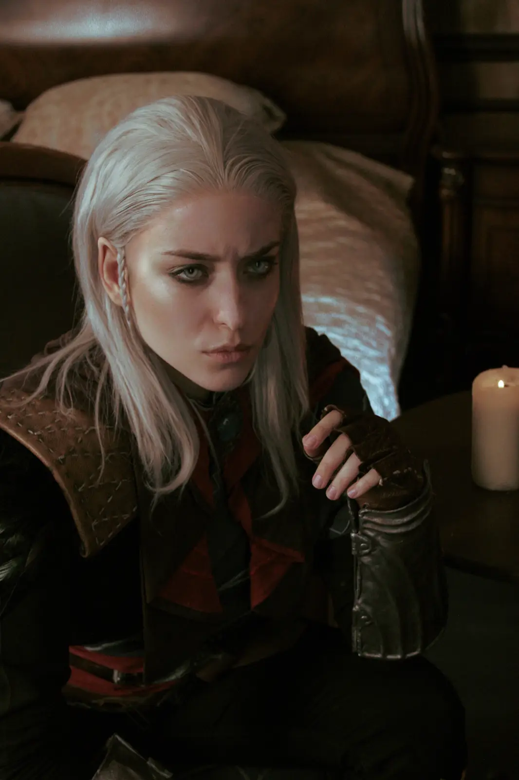 A woman with long silver hair and intense green eyes sits in a dimly lit room, wearing dark leather armor with red accents…