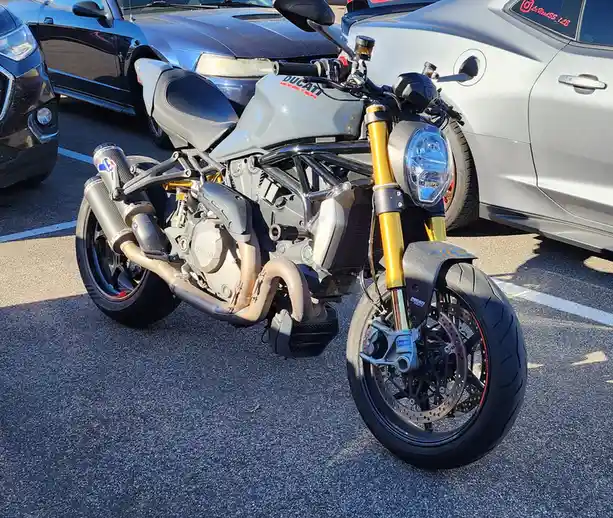 A gray Ducati Monster motorcycle with gold front forks is parked in a lot next to cars, with the overlaid text 'ducati…