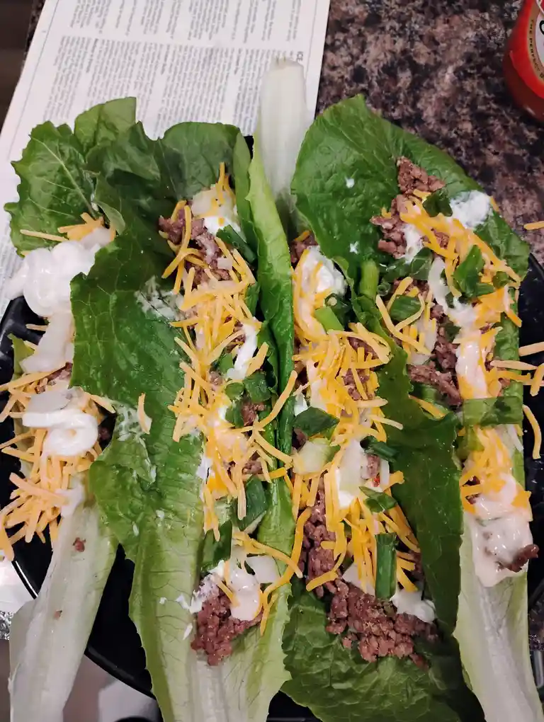 A close-up photo shows several lettuce wraps on a black plate, each filled with ground meat, shredded cheddar cheese…
