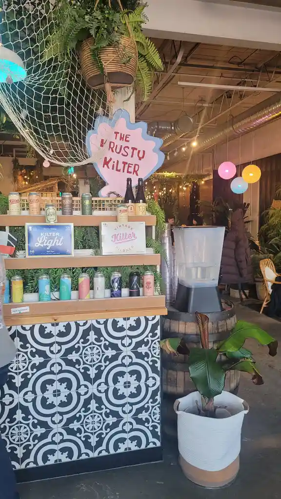 A photo of a beer stand called 'The Rusty Kilter' with shelves displaying various cans and bottles of beer, surrounded by…