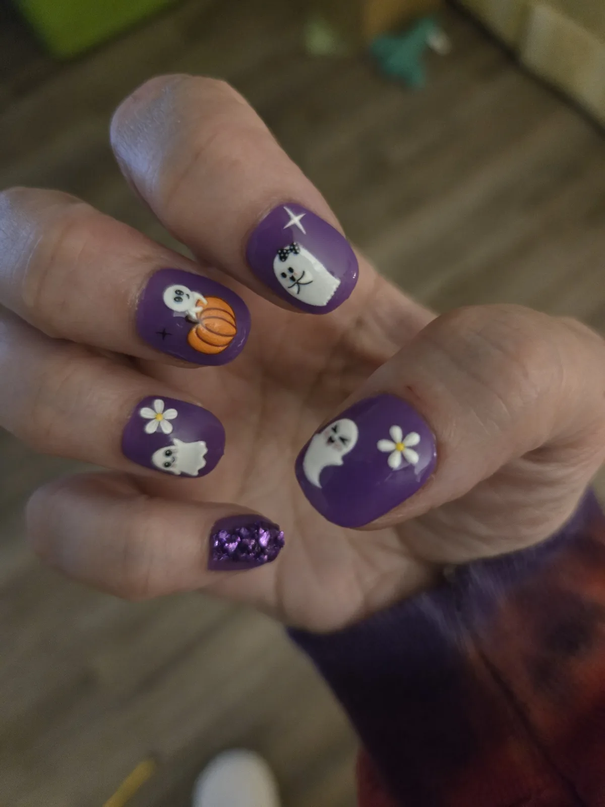 A close-up of a hand displaying Halloween-themed nail art on purple nails, featuring cute white ghosts, a pumpkin, daisies…