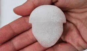 a close-up photo shows a hand holding a white, frost-covered, roughly spherical object shaped like a human head with a…