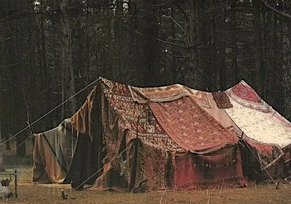 A makeshift tent in a forest, constructed entirely from draped and tied rugs and carpets of various patterns and colors…