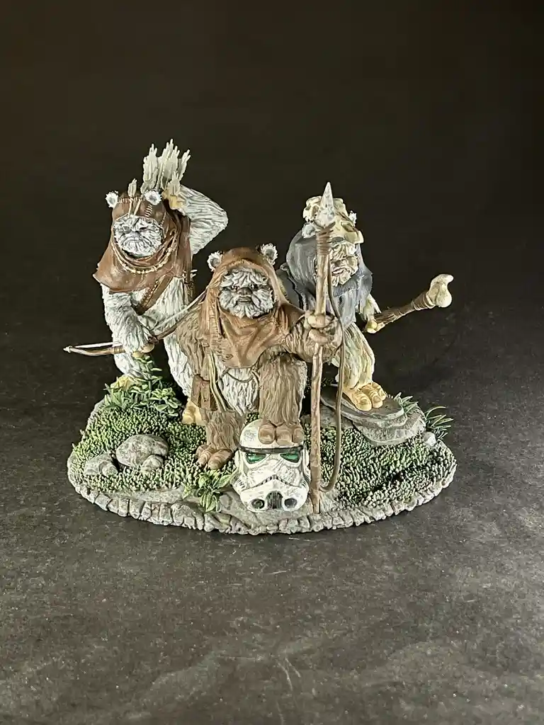 A painted miniature of three Ewoks stands on a mossy, rocky base against a dark background. The central Ewok, appearing as a…