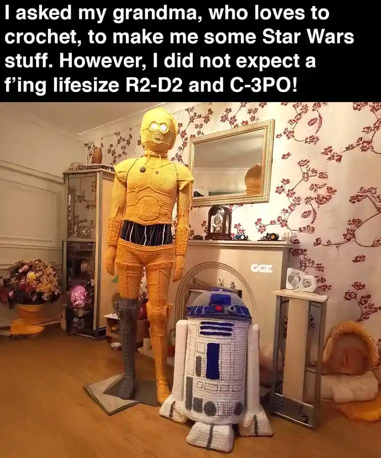 A room with floral wallpaper and wooden floors features two enormous, lifelike crocheted Star Wars droids: a full-sized…