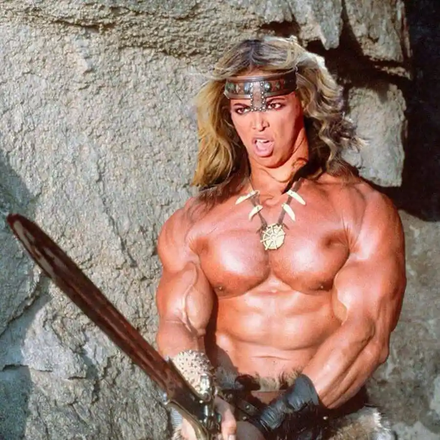 A muscular, shirtless man with long blonde hair, wearing a leather headband and tribal-style necklace, holds a sword while…