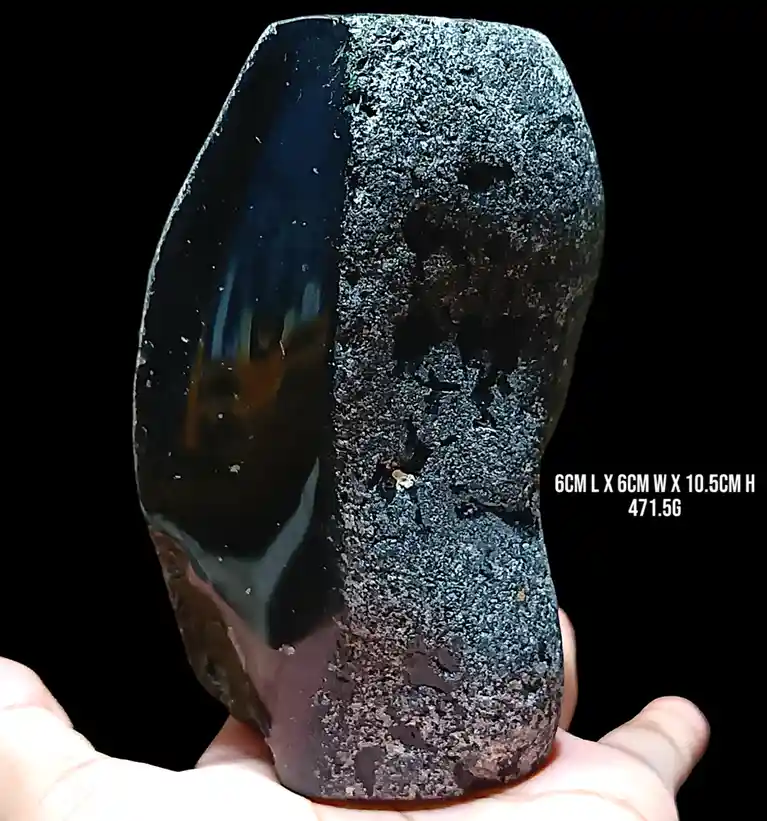 A hand holds a large, irregularly shaped black stone with one smooth, glossy side and one rough, textured side. White text…