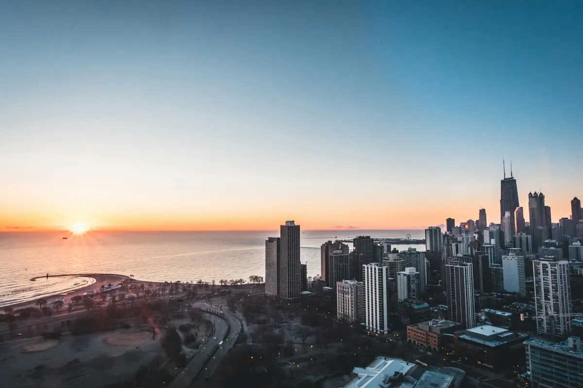 A scenic aerial photo of a city skyline at sunset, with the sun low on the horizon over water, casting orange and blue hues…