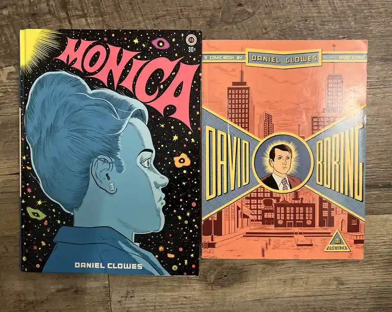 Two comic books by Daniel Clowes sit side by side on a wooden surface: 'Monica' with a blue-haired woman against a cosmic…