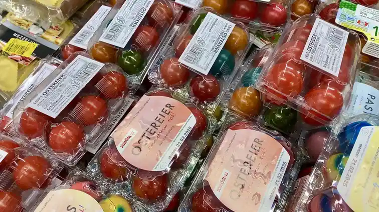 A supermarket shelf displays numerous clear plastic packages filled with colorful, dyed eggs labeled 'OSTEREIER' (German for…