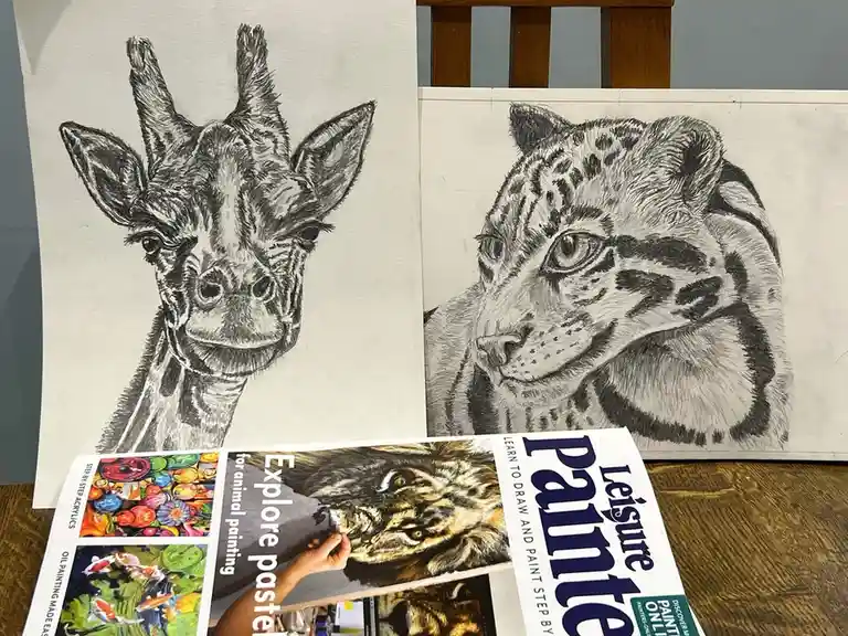 Two detailed black-and-white pencil drawings of a giraffe and a snow leopard are displayed on canvases, positioned above a…