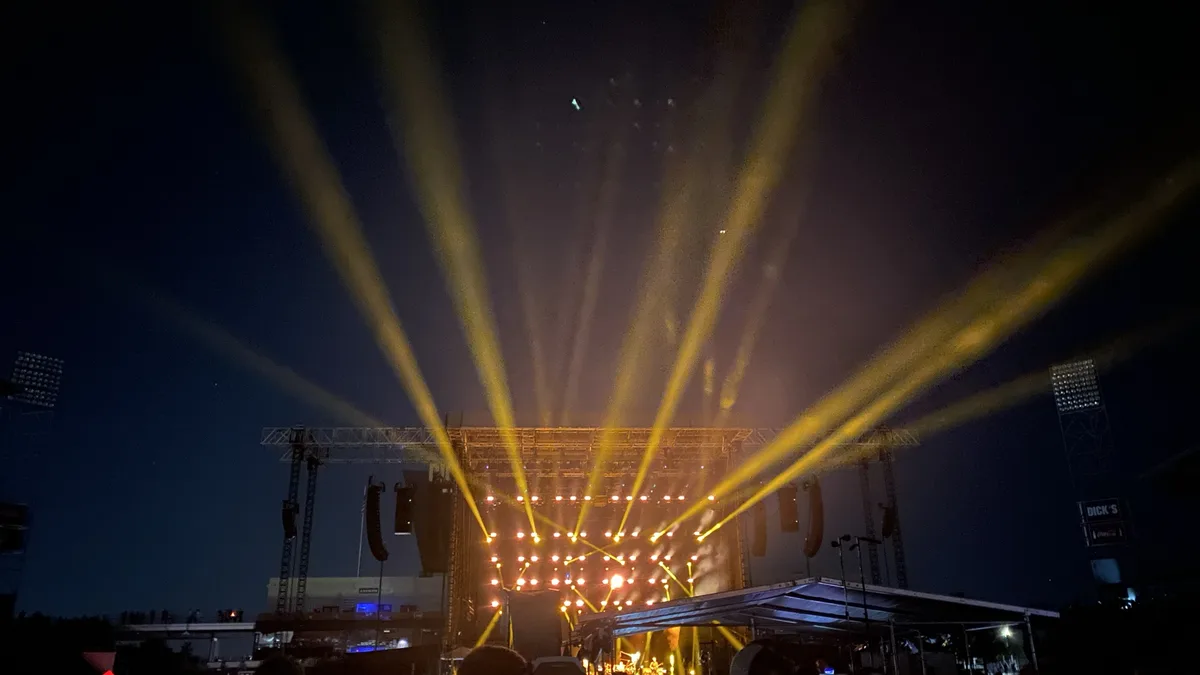 A nighttime concert scene with bright yellow stage lights beaming upward into the dark sky, illuminating haze. The stage…