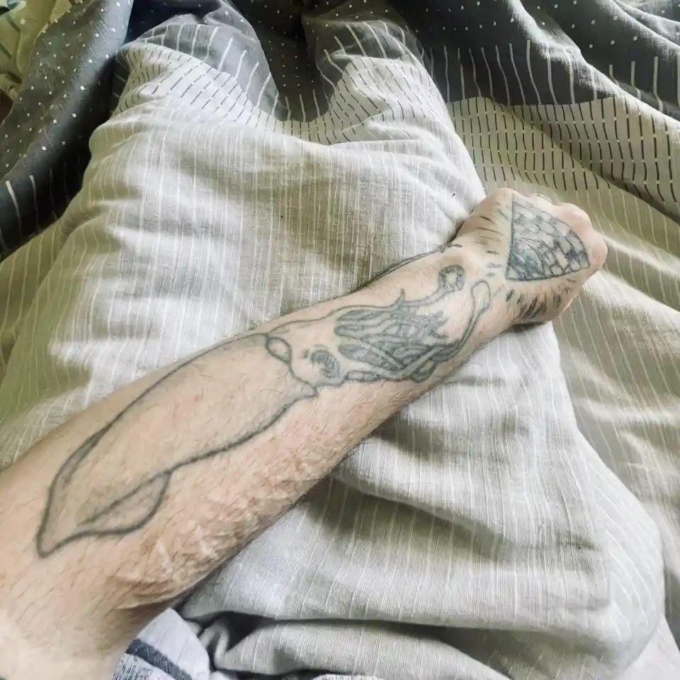 A close-up photo shows a person's tattooed forearm and clenched fist resting on a bed with patterned sheets. The tattoo is a…