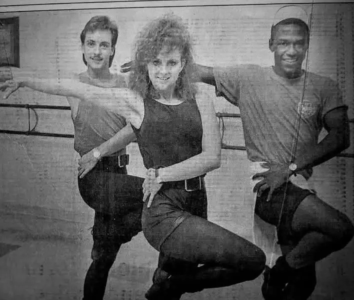 A grainy black-and-white photo shows three people in 1980s dance attire posing in a studio with barres in the background…