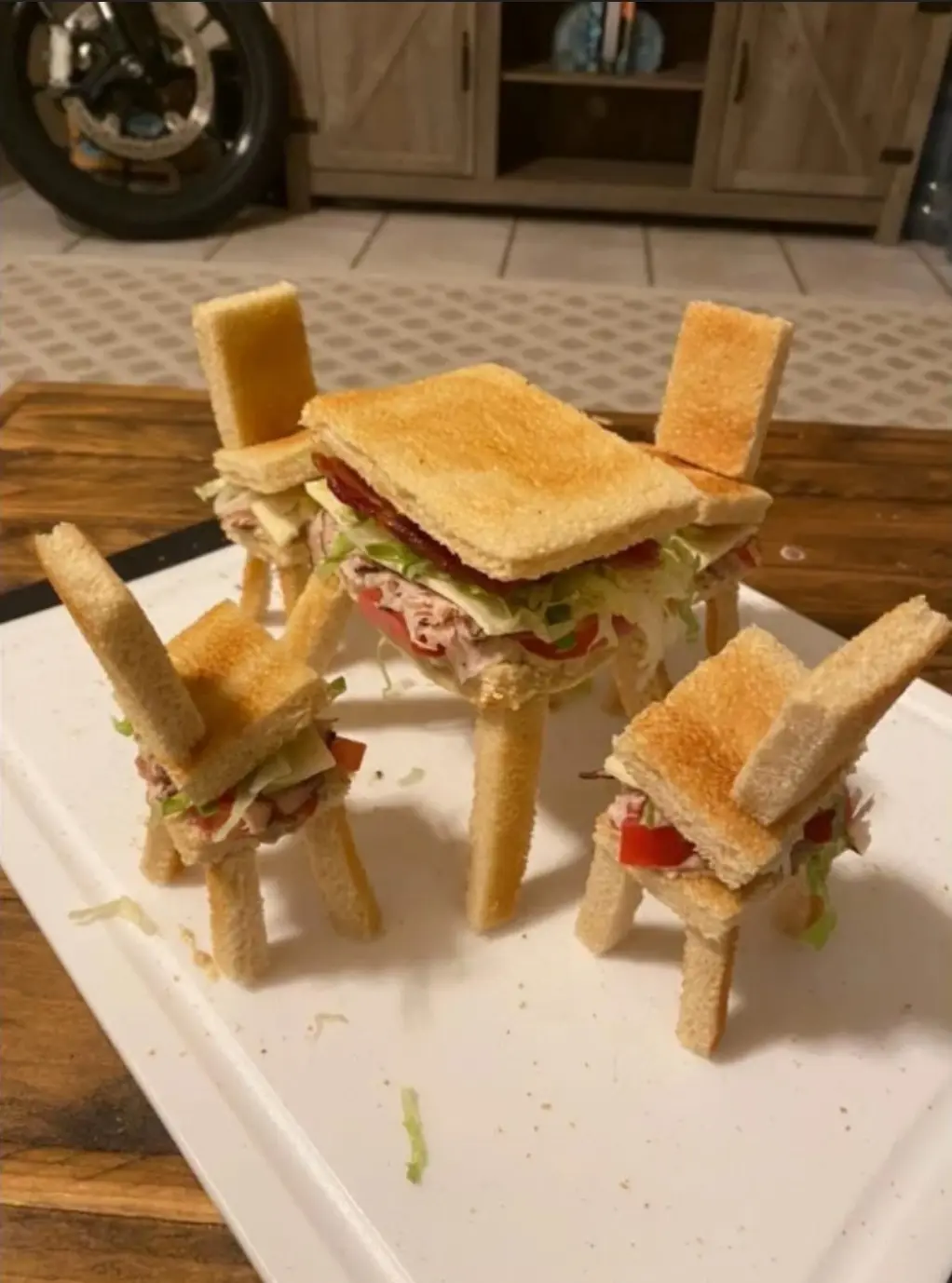 A white plate holds four tiny sandwiches crafted to resemble miniature chairs, each with a bread slice forming the seat and…