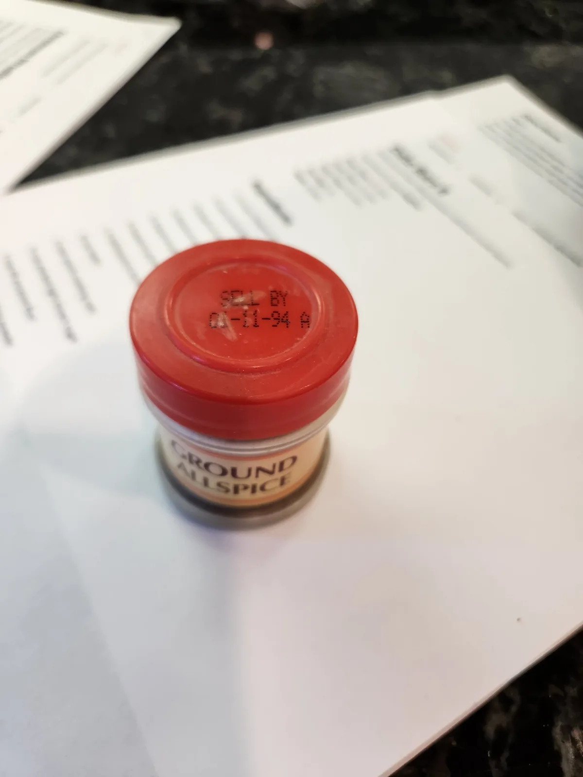 A small spice jar labeled 'GROUND ALLSPICE' sits on a white paper, its red lid showing a 'SELL BY 01-11-94' date, humorously…