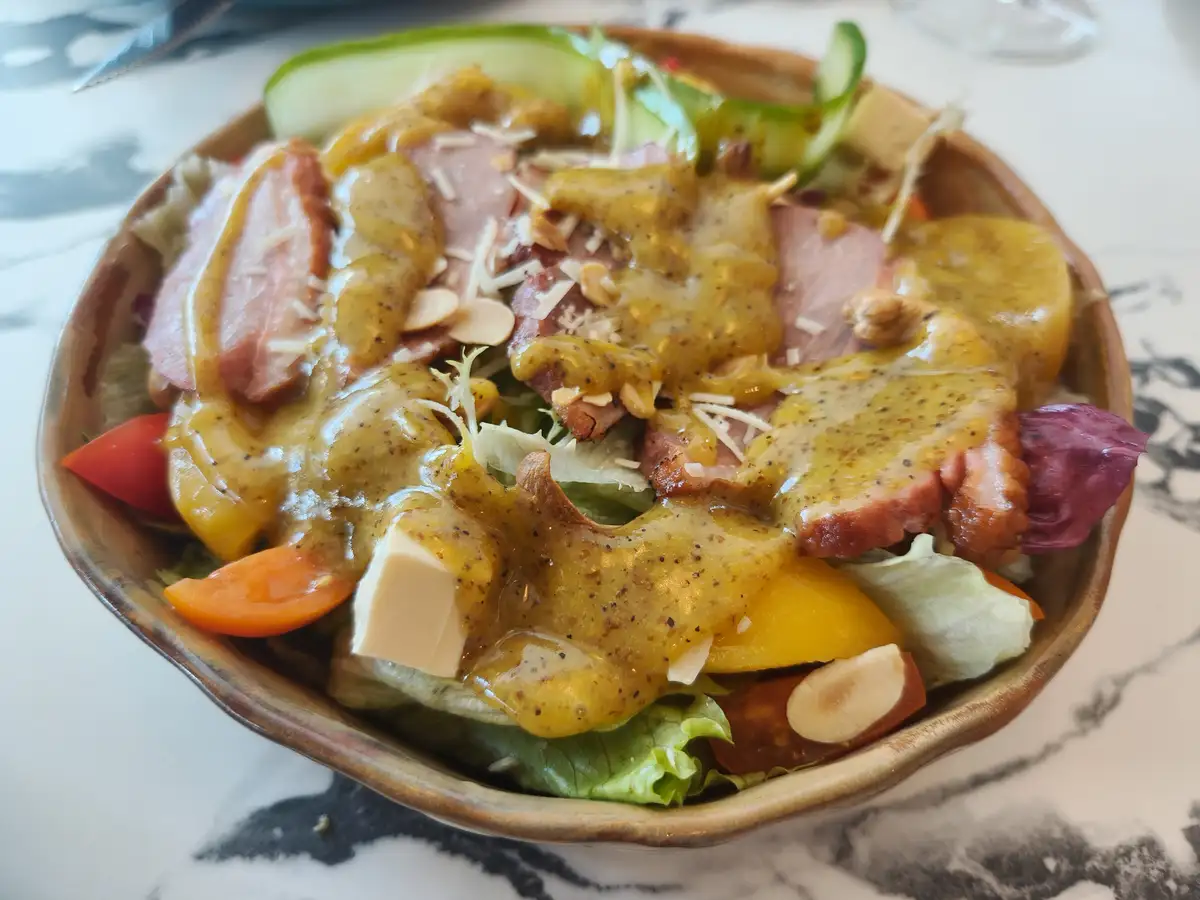 A close-up photo of a salad in a brown ceramic bowl, featuring lettuce, cucumber slices, tomato wedges, ham, cheese cubes…