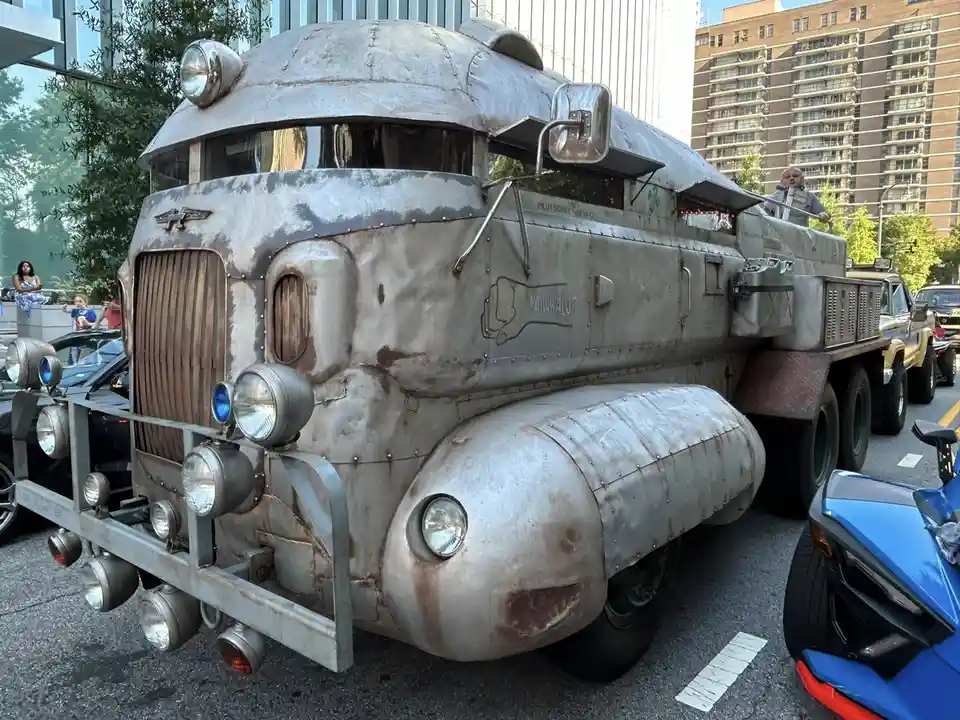 A heavily modified, custom-built, metallic, retro-futuristic truck with riveted panels, multiple headlights, and a bone logo…