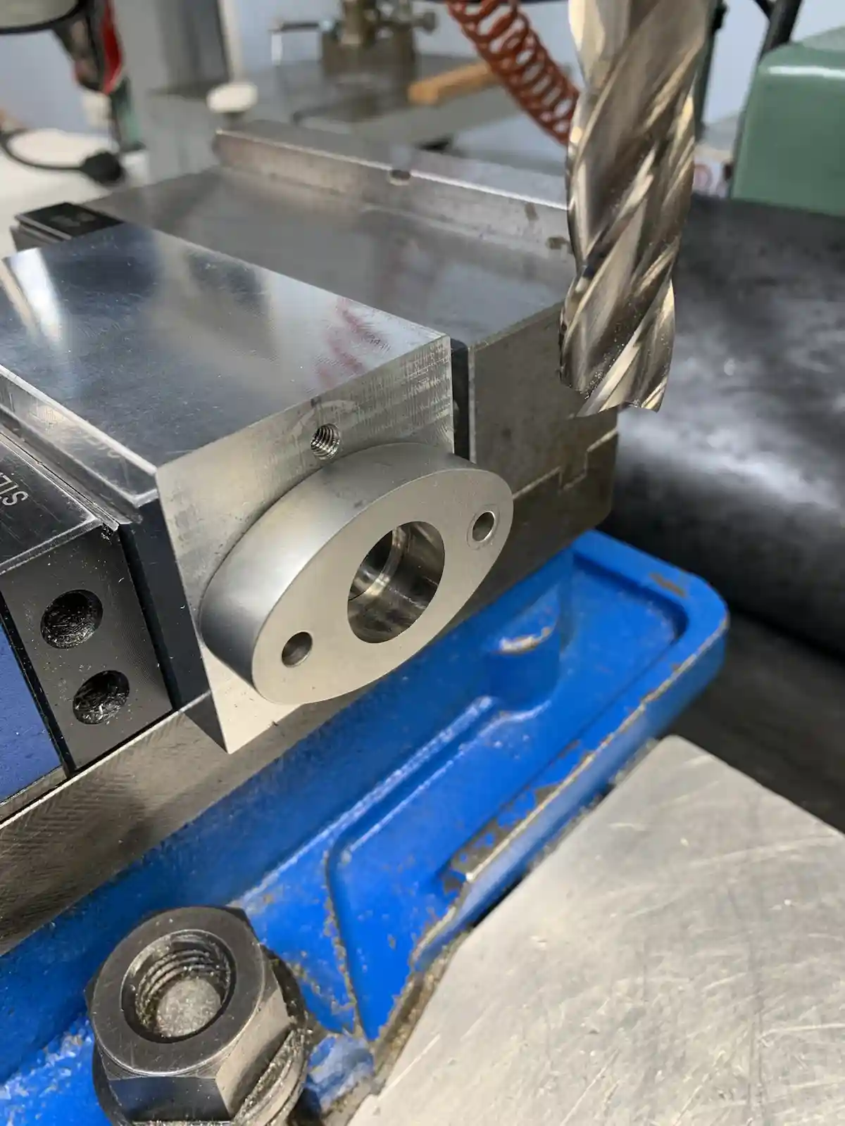 A close-up photo shows a metal drill bit hovering just above a machined metal part clamped in a blue vise. The part has a…