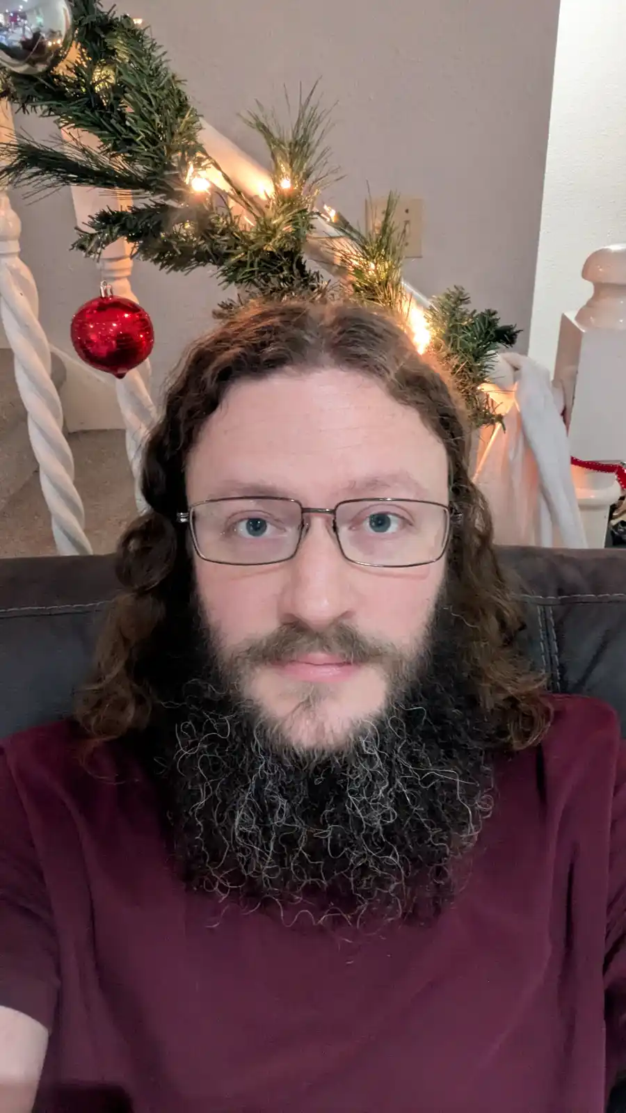 A man with glasses and a long beard sits on a couch, with a decorative Christmas tree branch and lights placed on his head…