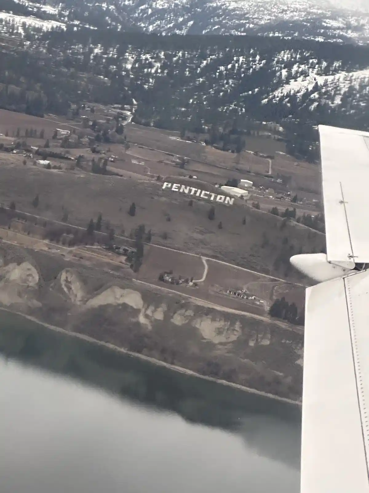 An aerial view from an airplane window shows the wing on the right, looking down at a landscape with snow-dusted mountains…