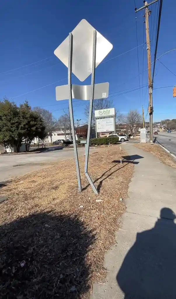 A photo of a bent, leaning metal signpost with two blank signs, standing on a grassy verge next to a sidewalk under a clear…