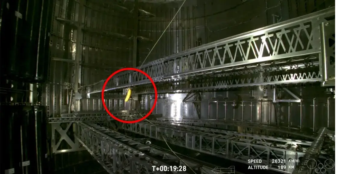 A still from a high-speed rocket launch video shows a single yellow banana hanging from a metal truss structure inside the…