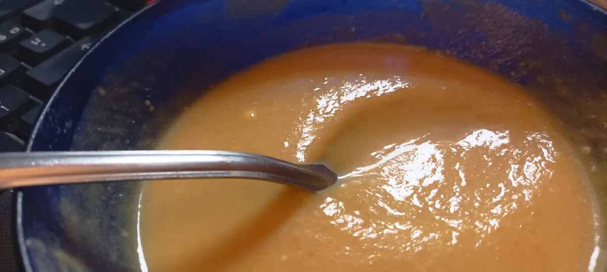 A close-up photo shows a thick, brownish-orange soup or sauce in a dark blue bowl, with a metal spoon resting in it. Part of…