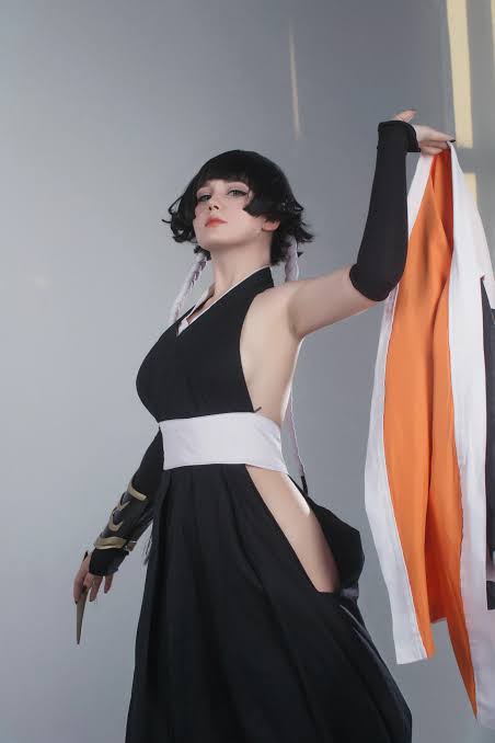 A person in a black and white cosplay outfit, holding up an orange and white fabric, looks upward with a displeased…