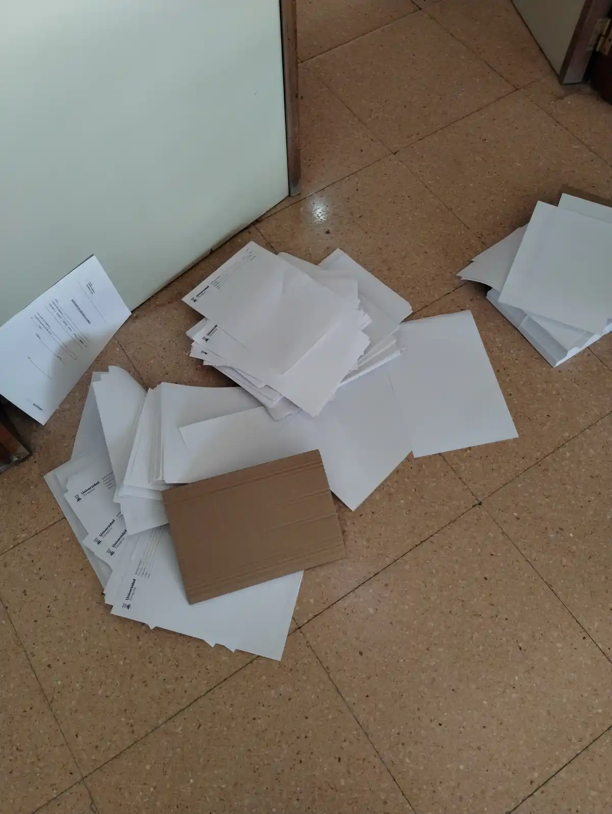 A high-angle photo shows numerous white envelopes and papers scattered across a tiled floor, with one brown cardboard piece…