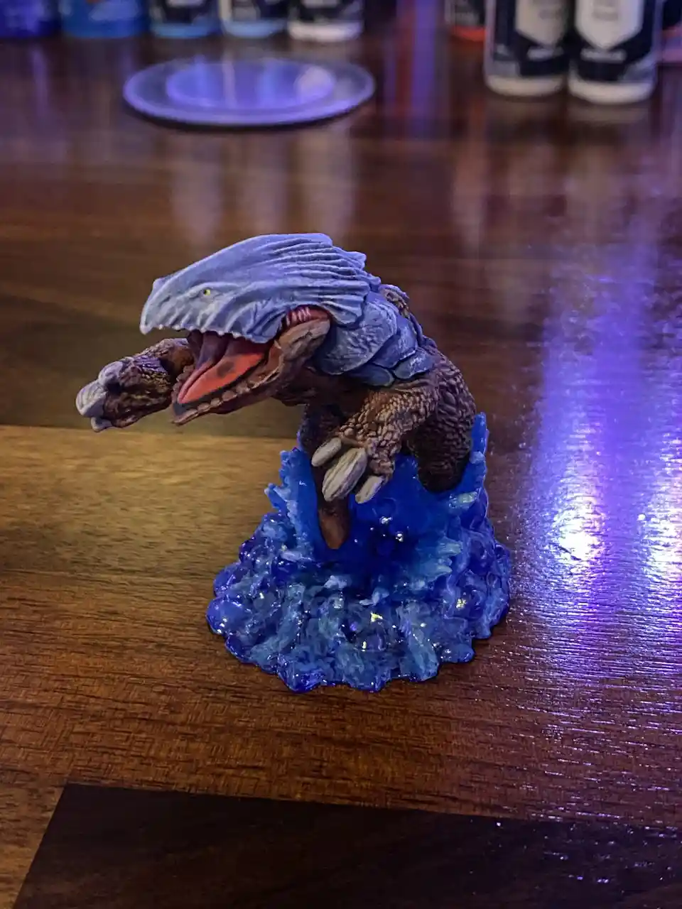 A painted miniature monster figurine with a blue, textured head and brown body, standing on a blue, crystalline base on a…