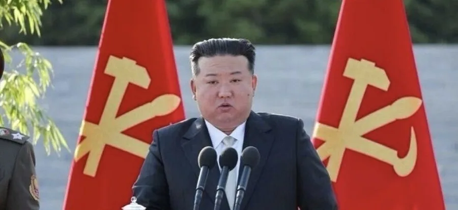 A man in a dark suit stands at a podium with three microphones, flanked by two red flags bearing golden hammer and sickle…