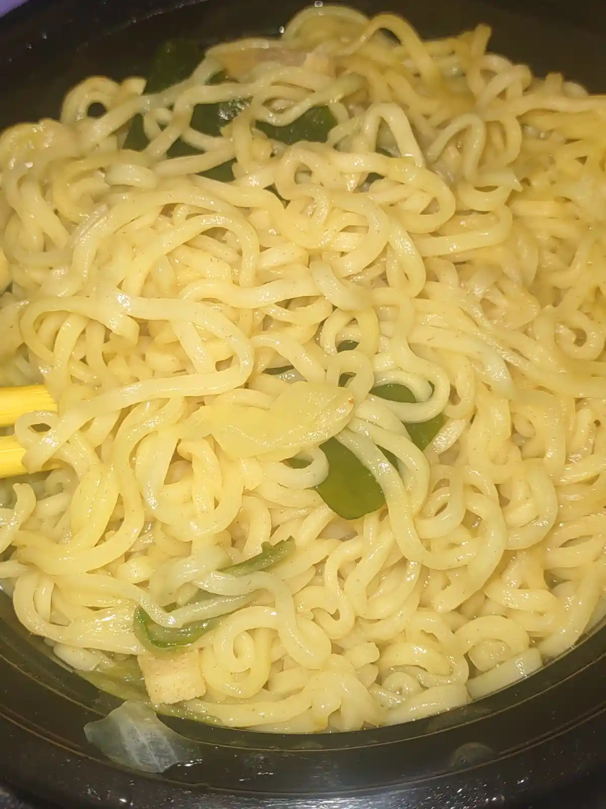 A close-up photo shows a bowl of cooked ramen noodles with green vegetables and a piece of tofu, being stirred with yellow…