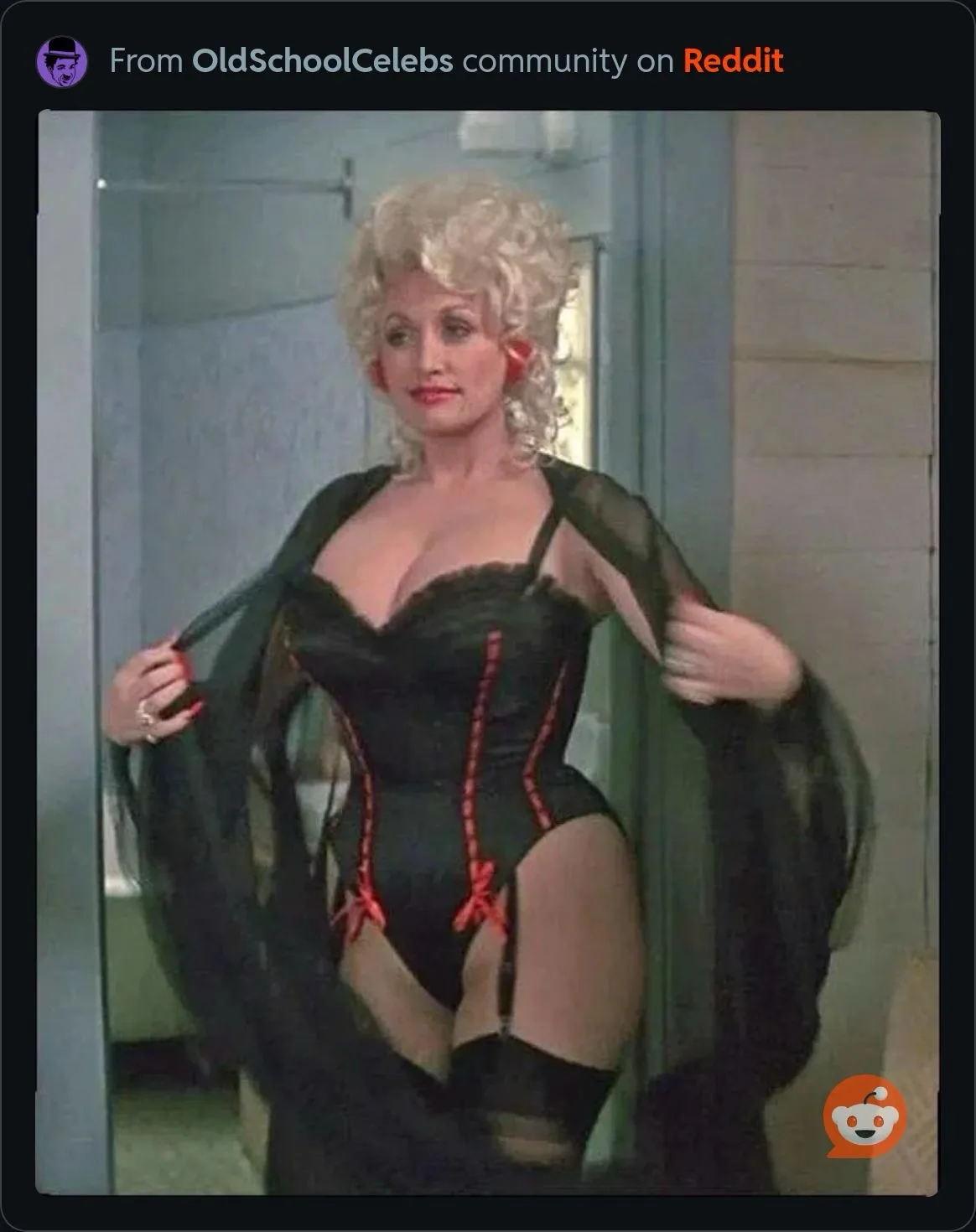 A woman with blonde curly hair and red lipstick stands in a bathroom, pulling open a sheer black robe to reveal a black…