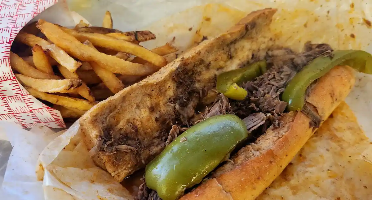 A close-up photo shows a partially eaten sandwich filled with shredded meat and green peppers, served alongside a side of…