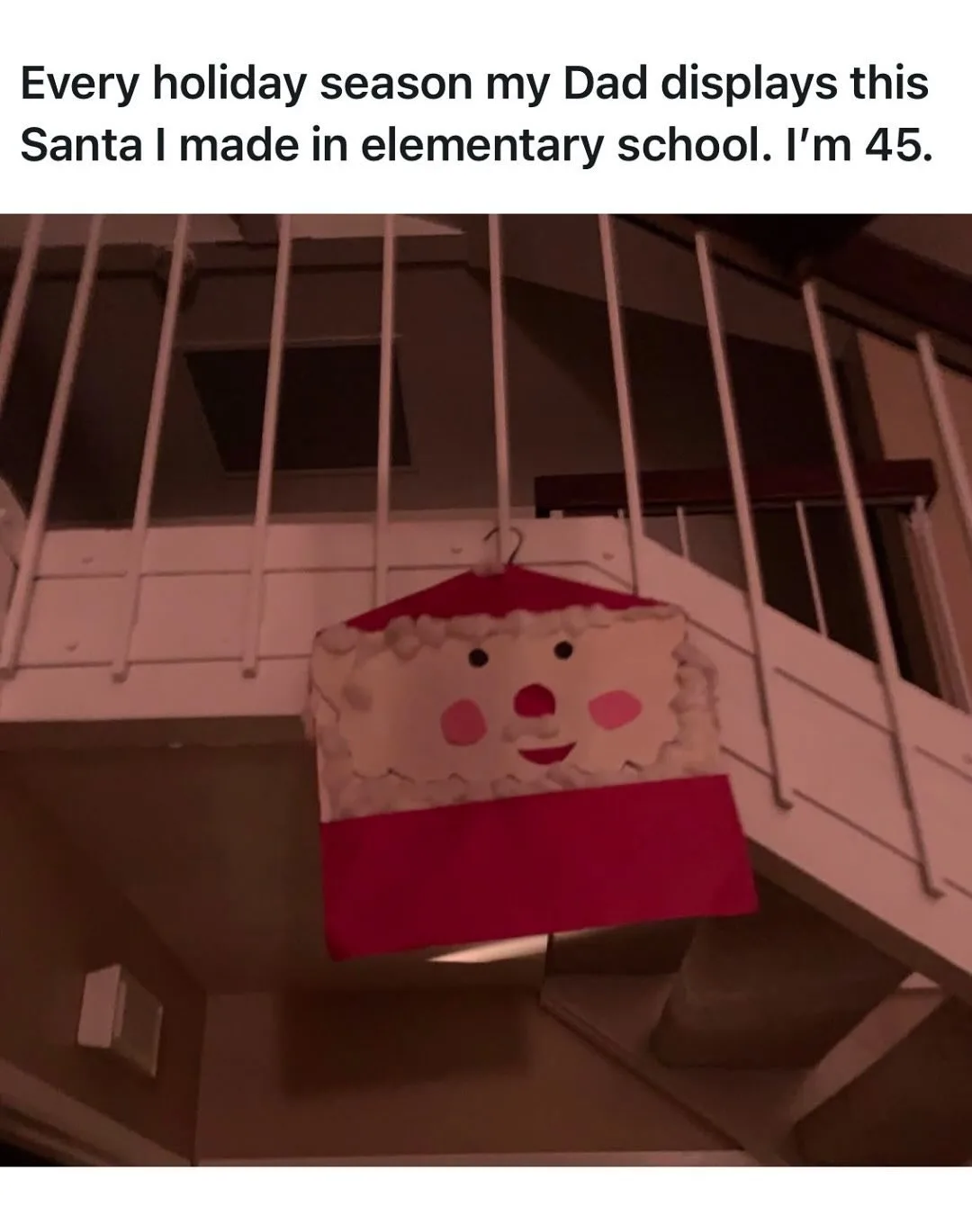 A handmade, childlike Santa Claus decoration made of paper and fabric hangs from a white staircase railing. The Santa has a…