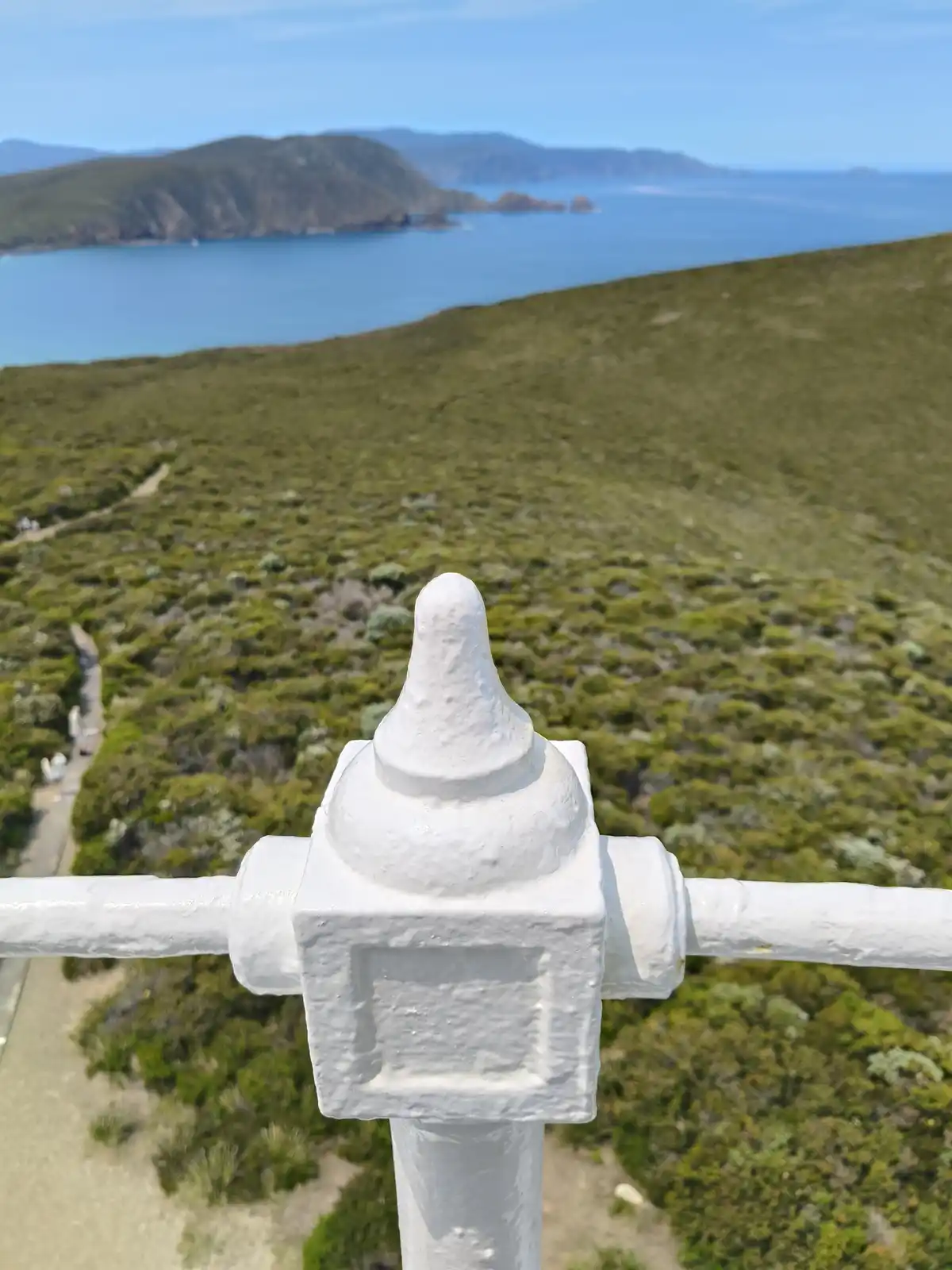 A close-up photo shows the top of a white metal railing post with a rounded finial, overlooking a scenic coastal landscape…