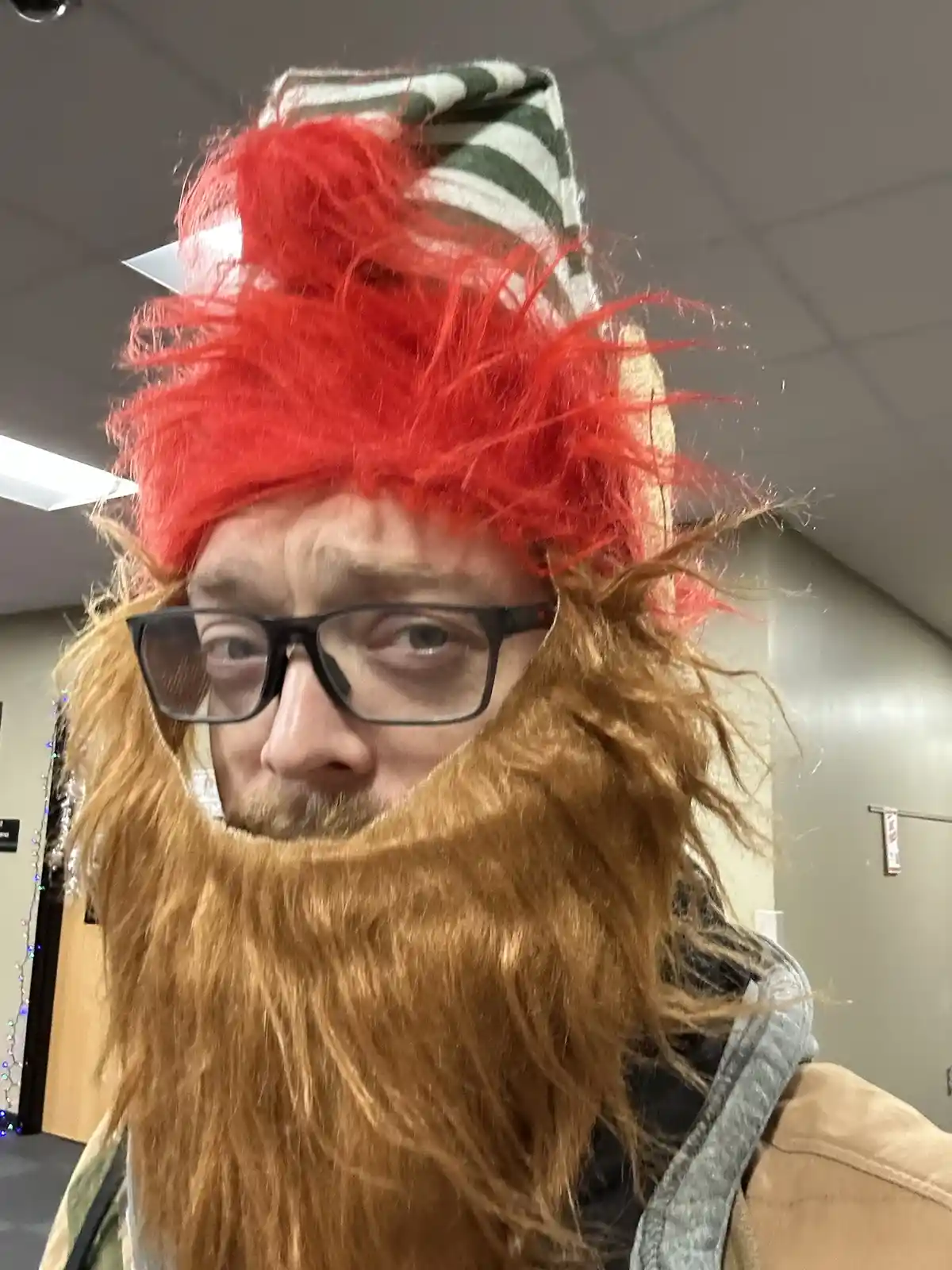 A man wearing glasses and a messy red wig, with a large brown beard covering his lower face, also wearing a green and white…