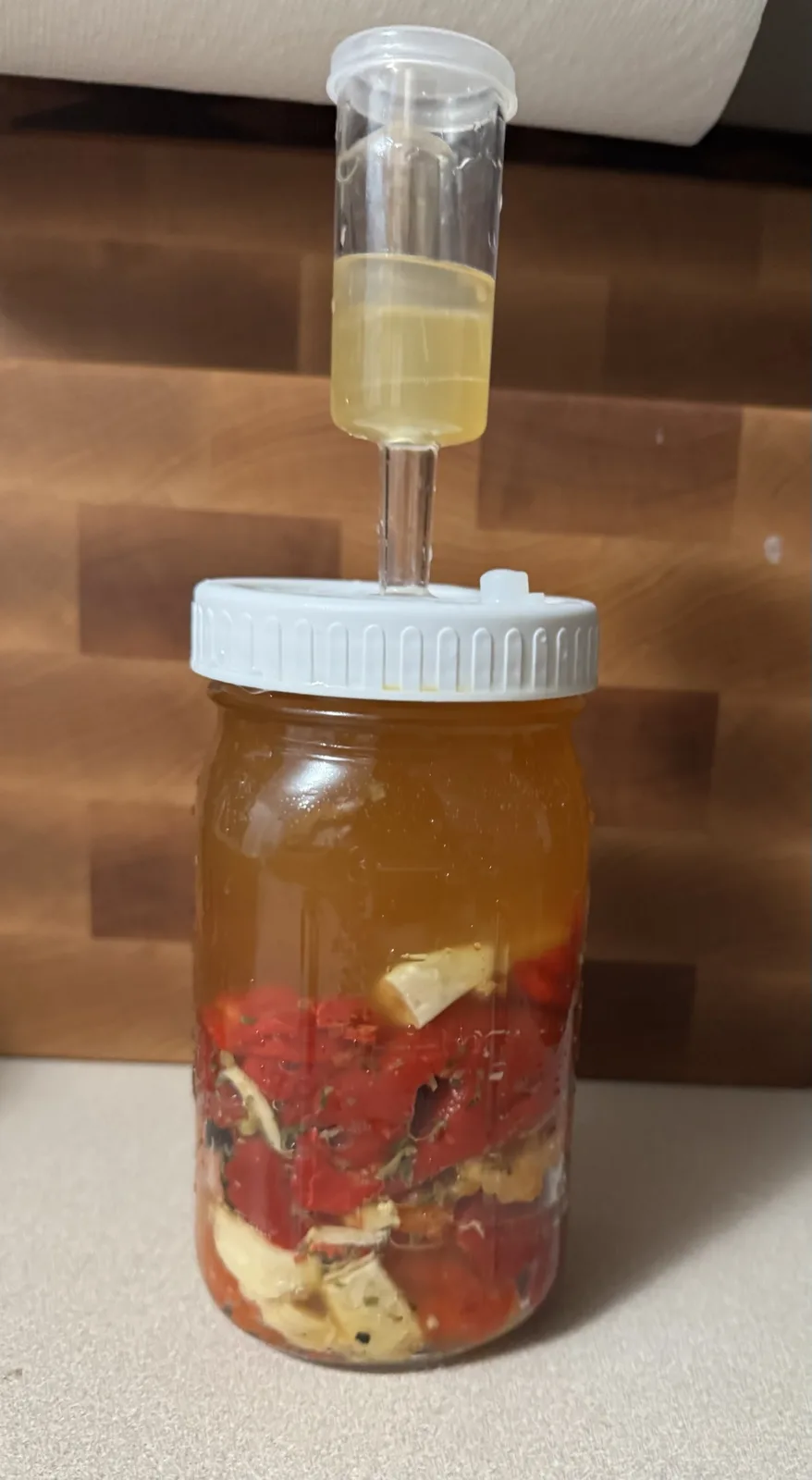 A glass mason jar filled with chopped red tomatoes and white cheese pieces submerged in a golden liquid, topped with a white…