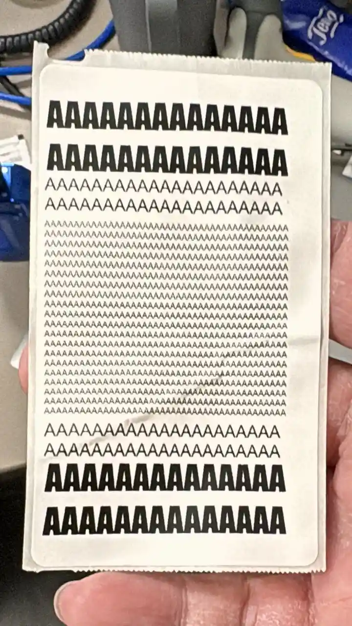 A hand holds a white rectangular card with multiple rows of the letter 'A' printed in bold black font, varying in length and…