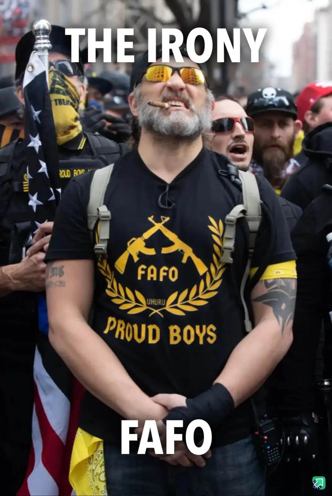 A man with a gray beard and yellow-tinted sunglasses stands in a crowd, wearing a black t-shirt with a yellow emblem…