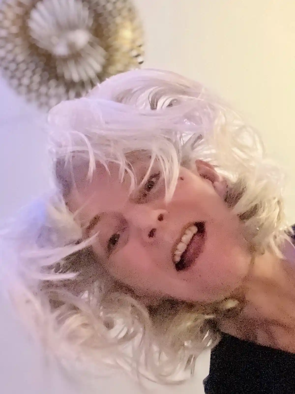 A person with voluminous, messy white hair is captured in a tilted, close-up selfie, their face contorted in an exaggerated…