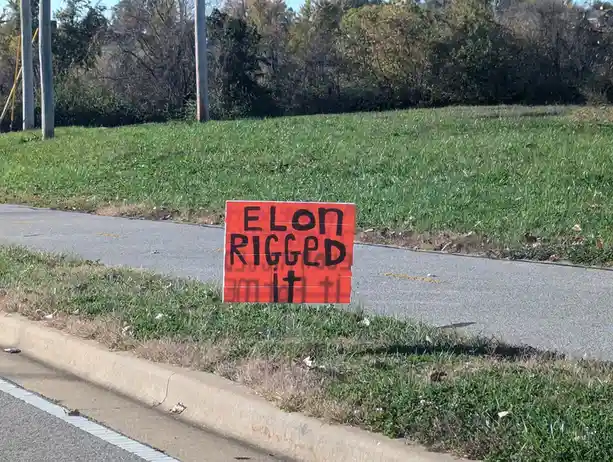 A bright orange protest sign with bold black handwritten text reading 'ELON RIGGED it' is stuck in grass beside a paved…