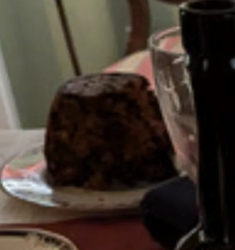a blurry photo shows a dark, dense, round fruitcake-like dessert on a plate, likely a christmas pudding, next to a glass and…