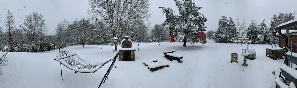 A wide snowy backyard panorama shows a snow-covered hammock, outdoor pizza oven, benches, and garden ornaments all blanketed…
