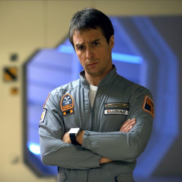 A man in a grey 'LUNAR TECHNICAL MAINTENANCE CREW' uniform stands with arms crossed, looking skeptical and unimpressed. The…