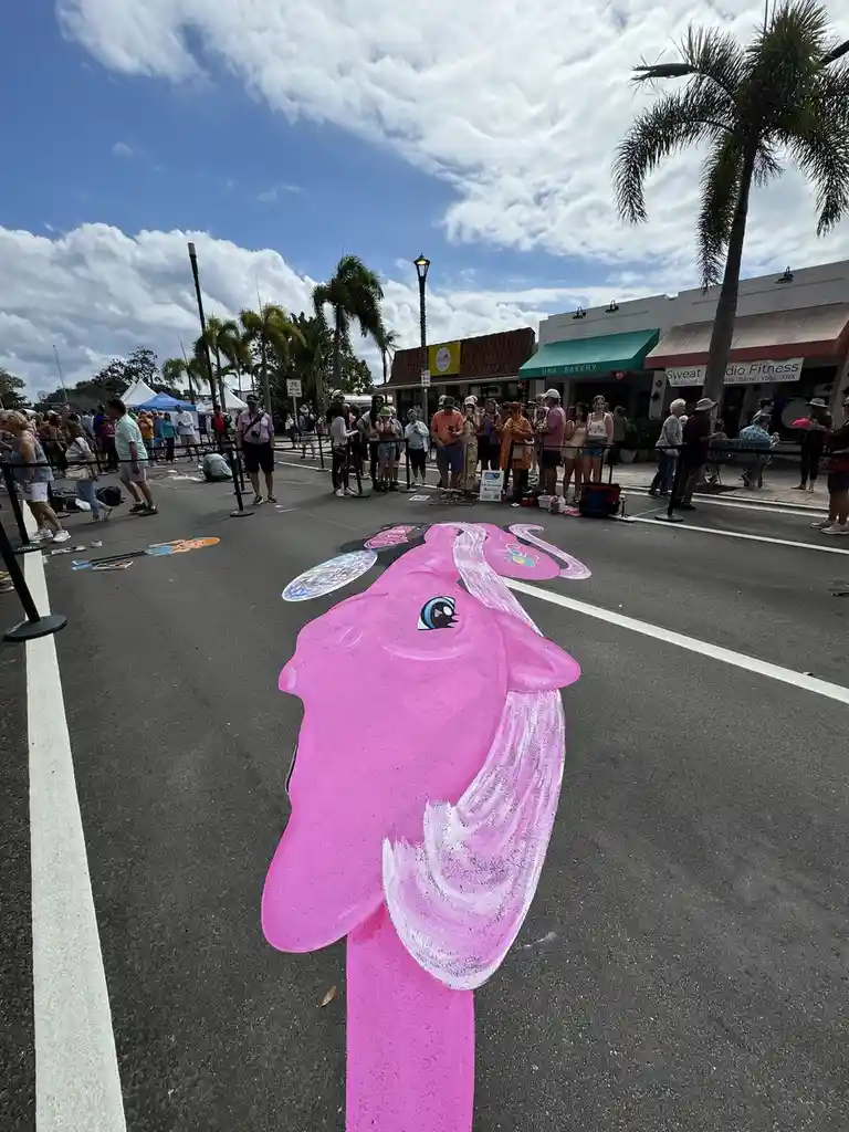 A large, vibrant pink chalk drawing of a pony's head and neck is on a paved street, with crowds of people standing behind…