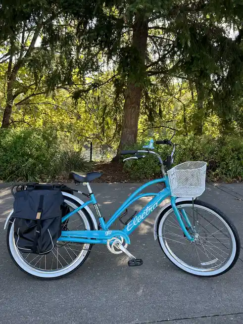 A bright blue Electra bicycle with a white basket on the front and a black pannier bag on the rear rack is parked on a paved…